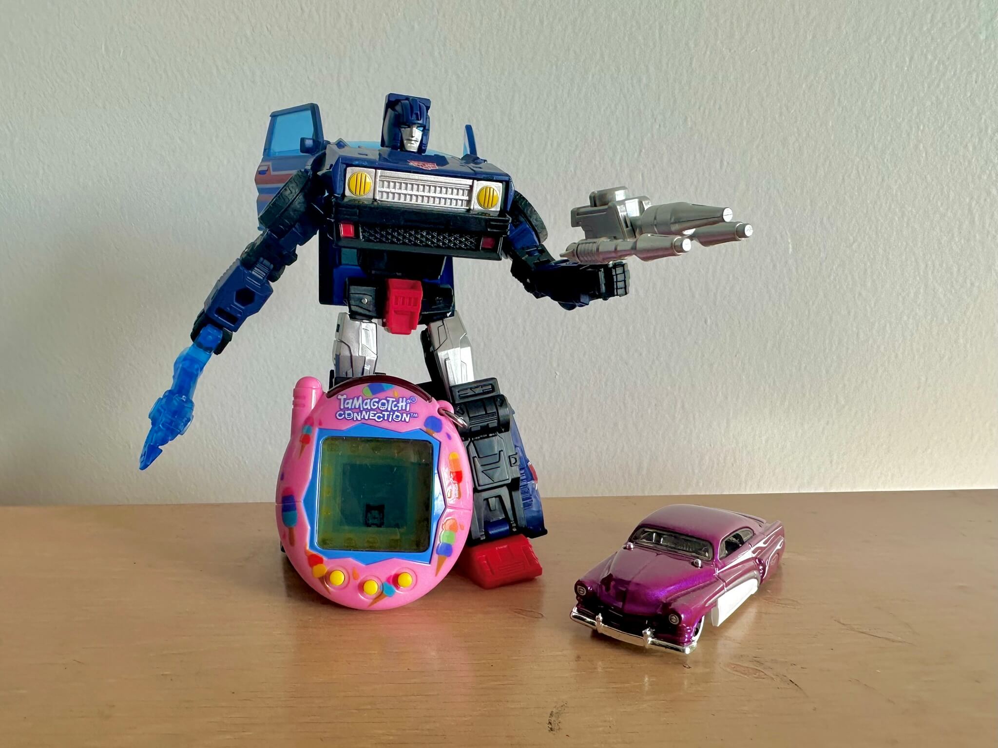 Transformer in robot mode with pink Tamagotchi leaning against its legs, and a Hot Wheels car next to it
