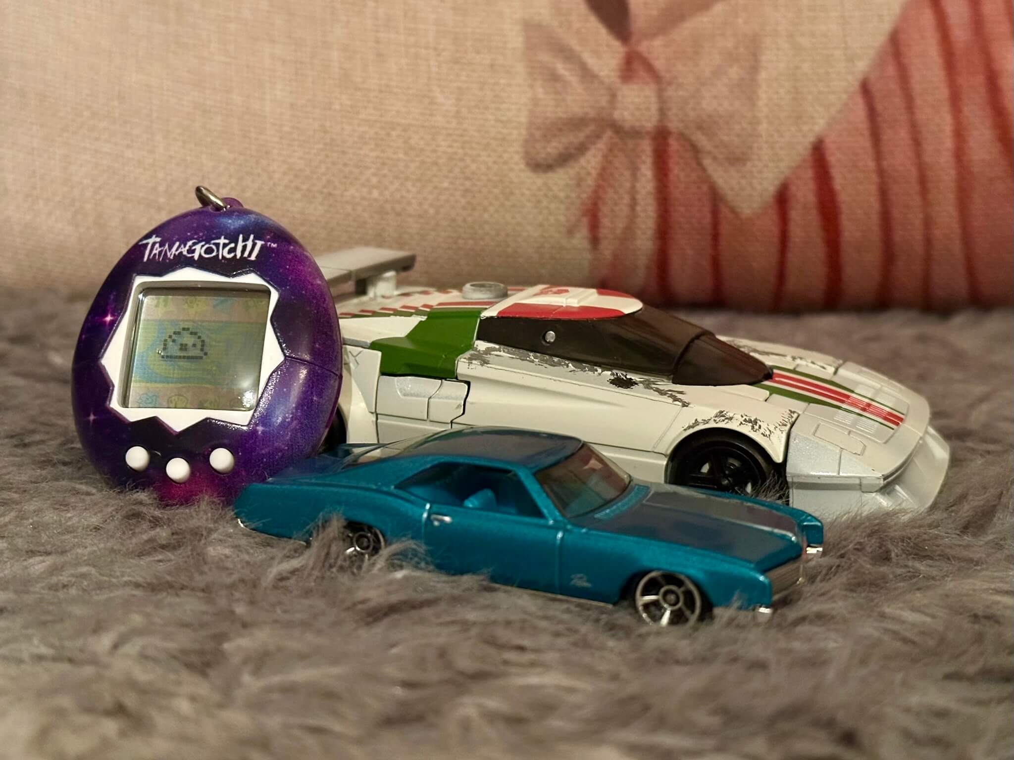 A large white car and a small blue-green car with a purple Tamagotchi resting on them, sitting on a fluffy surface