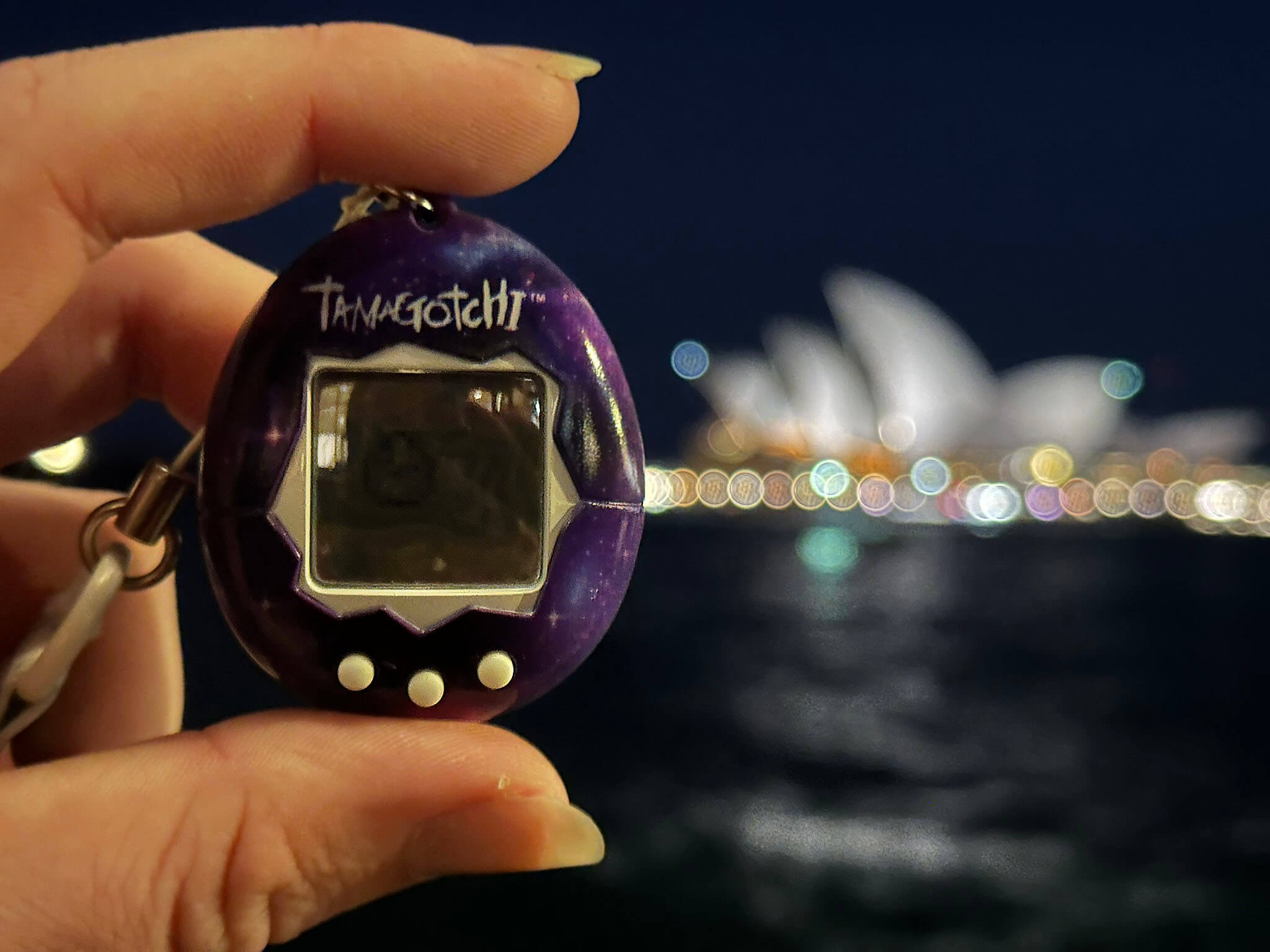 Purple Tamagotchi held up with the Sydney Opera House blurred in the background