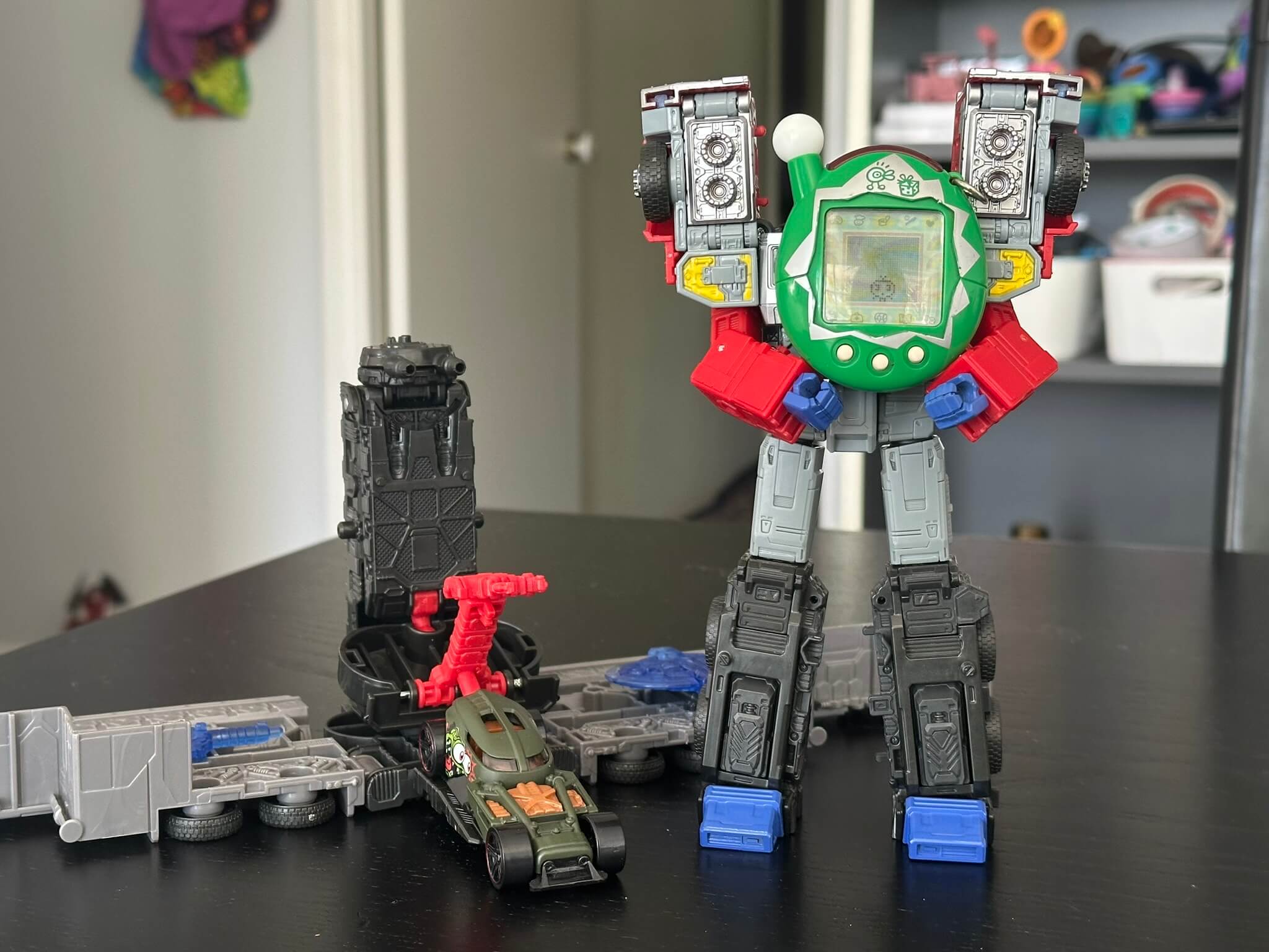 Transformer holding a green Tamagotchi, and a Hot Wheels car parked in the repair bay of the Transformers base mode