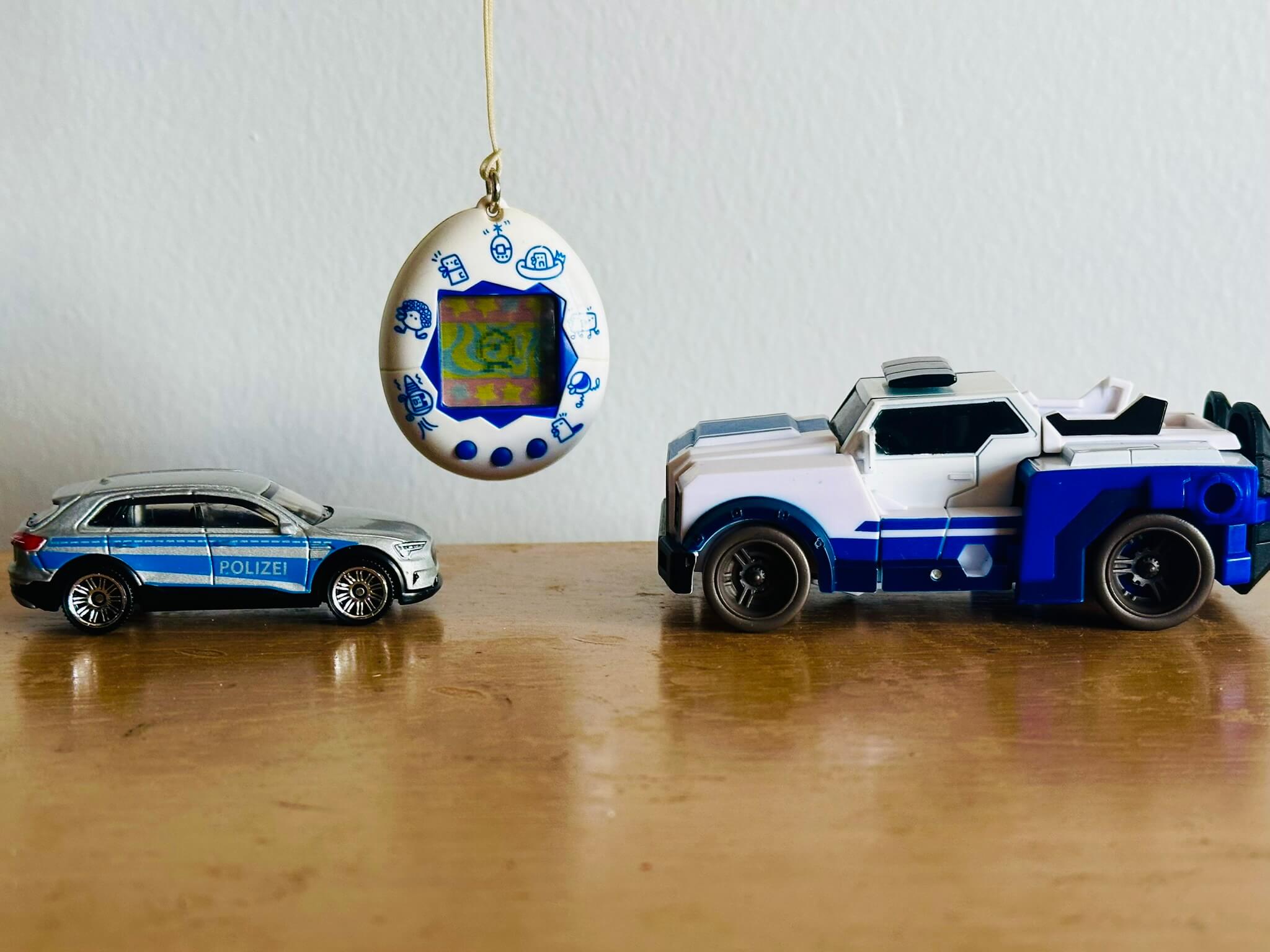 Silver toy car, white and blue Transformer and a white Tamagotchi suspended in the air between them