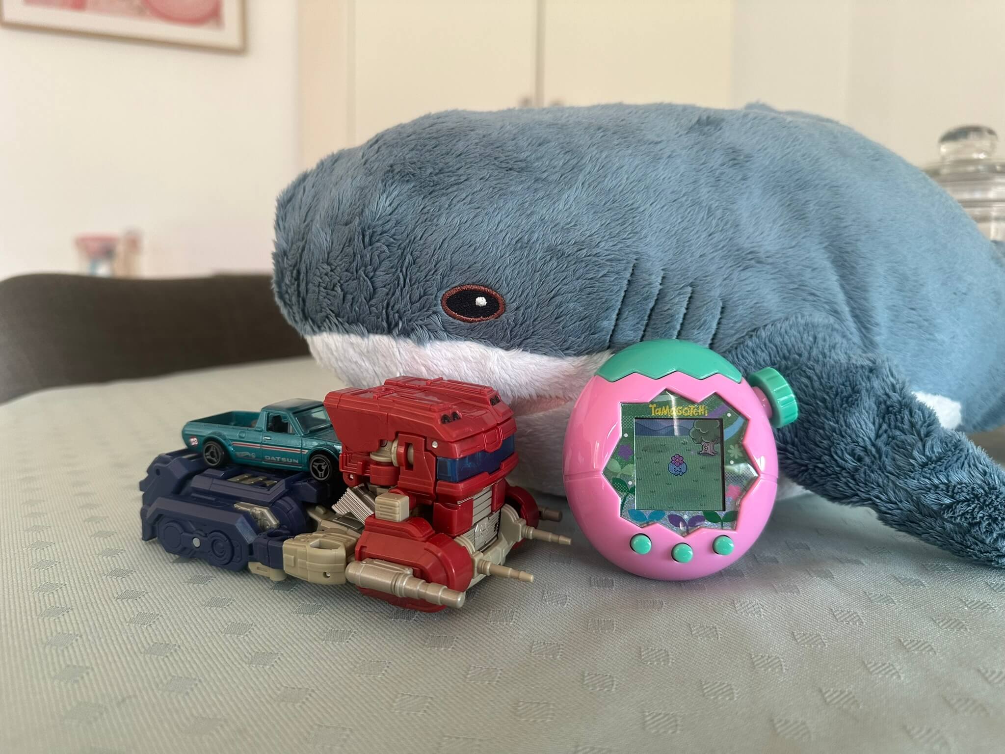 Pink Tamagotchi leaning against shark plushie, red truck Transformer with teal toy car on its back